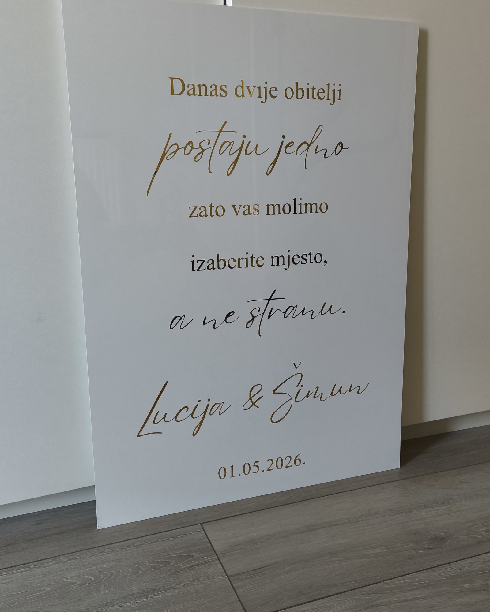 Bijela akrilna ploča s porukom „Danas dvije obitelji postaju jedno, zato vas molimo izaberite mjesto, a ne stranu“ i imenima Lucija & Šimun, 1. 5. 2026.
