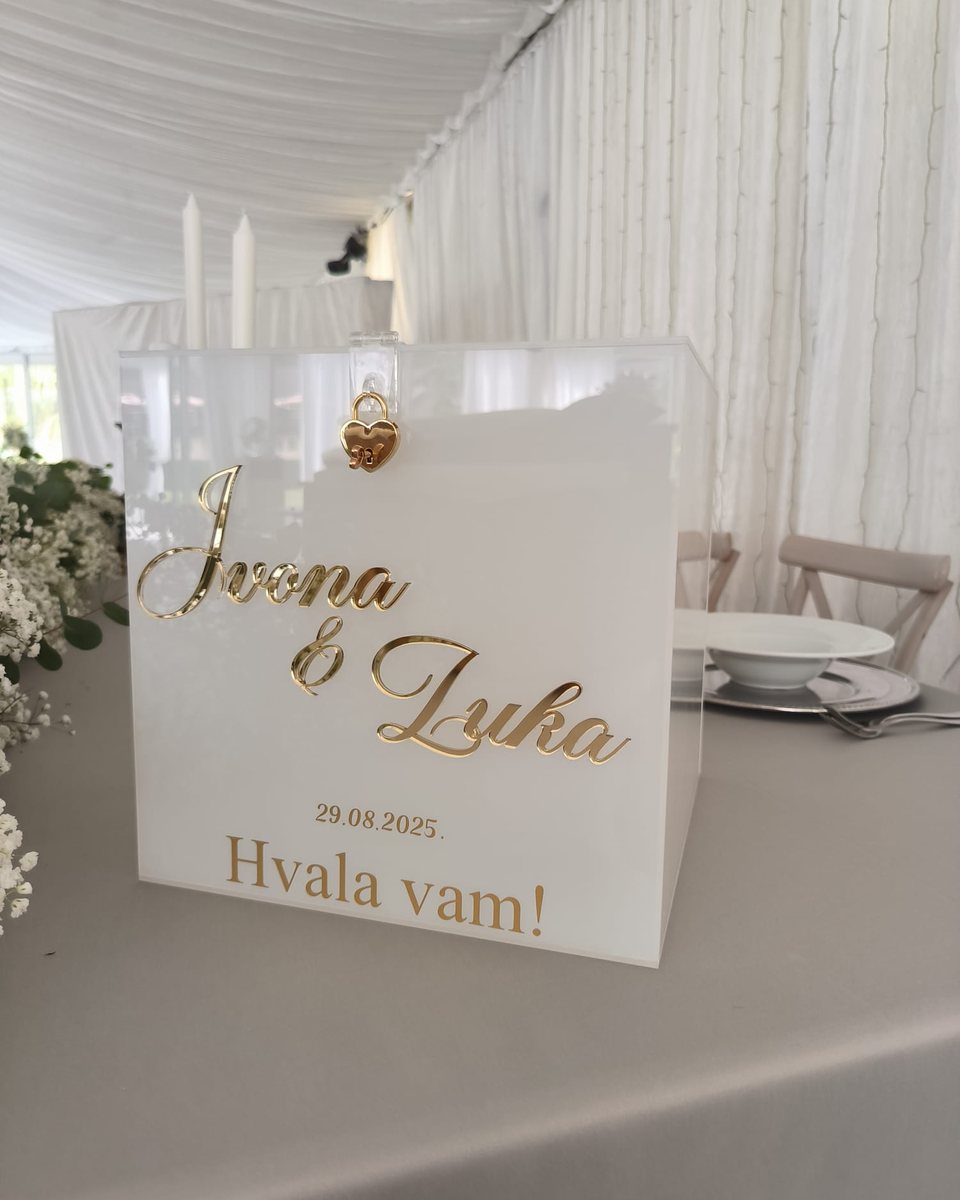 Bijela akrilna kutija za kuverte sa zlatnim natpisom „Ivona & Luka, 29. 8. 2025., Hvala vam!“ na stolu u svadbenom šatoru.