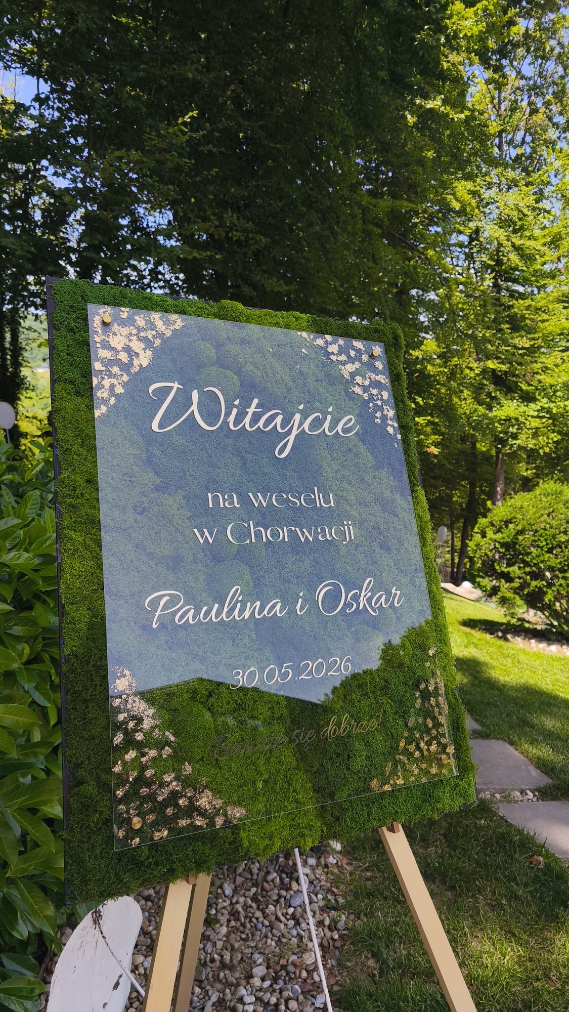 Welcome ploča s okvirom od mahovine i poljskim natpisom „Witajcie na weselu w Chorwacji, Paulina i Oskar“, na stalku na travnjaku uz drveće.
