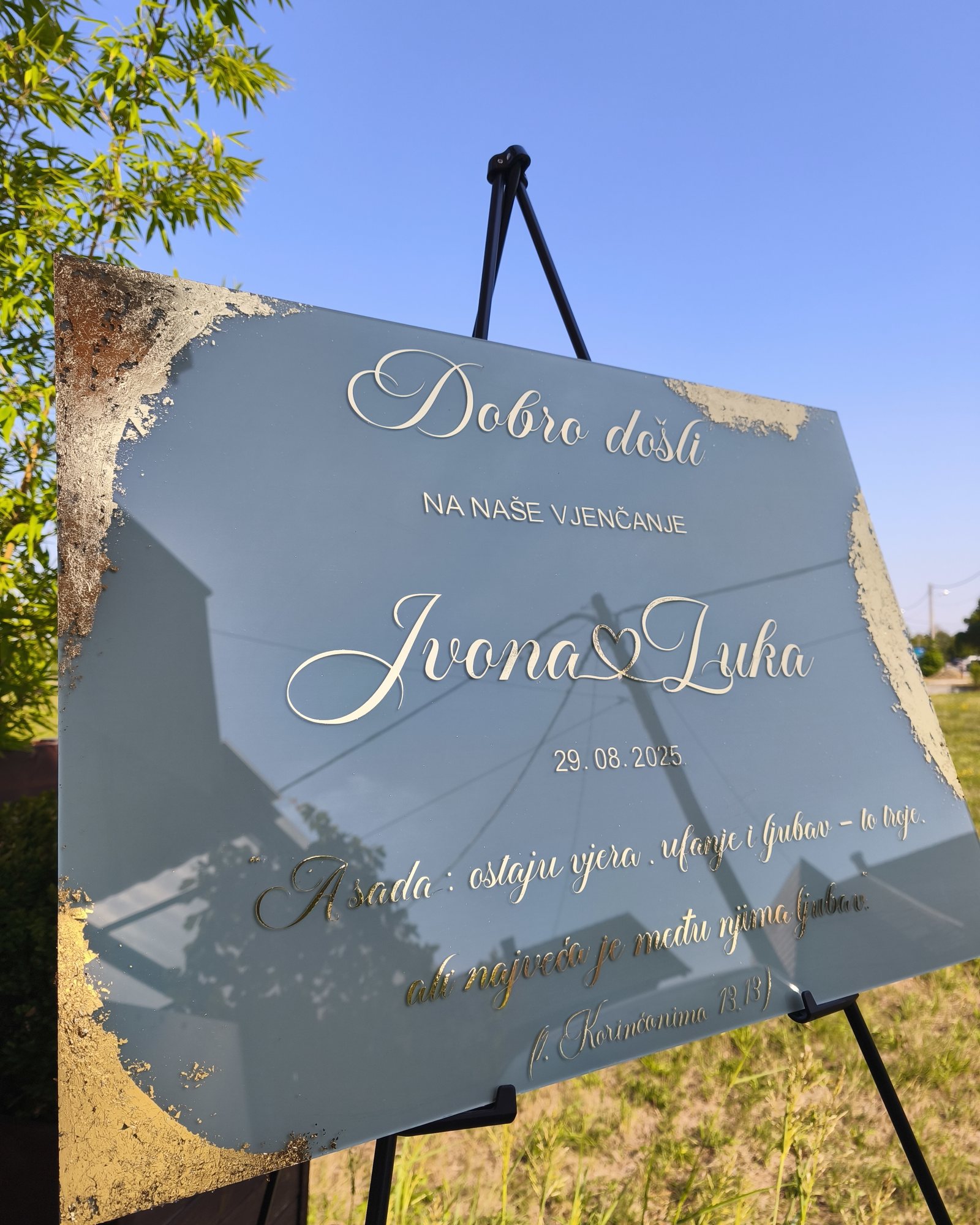 Ogledalo dobrodošlice sa zlatnim rubovima na crnom stalku na livadi, natpis „Dobro došli na naše vjenčanje, Ivona i Luka, 29. 8. 2025.“ i citat iz Prve poslanice Korinćanima.