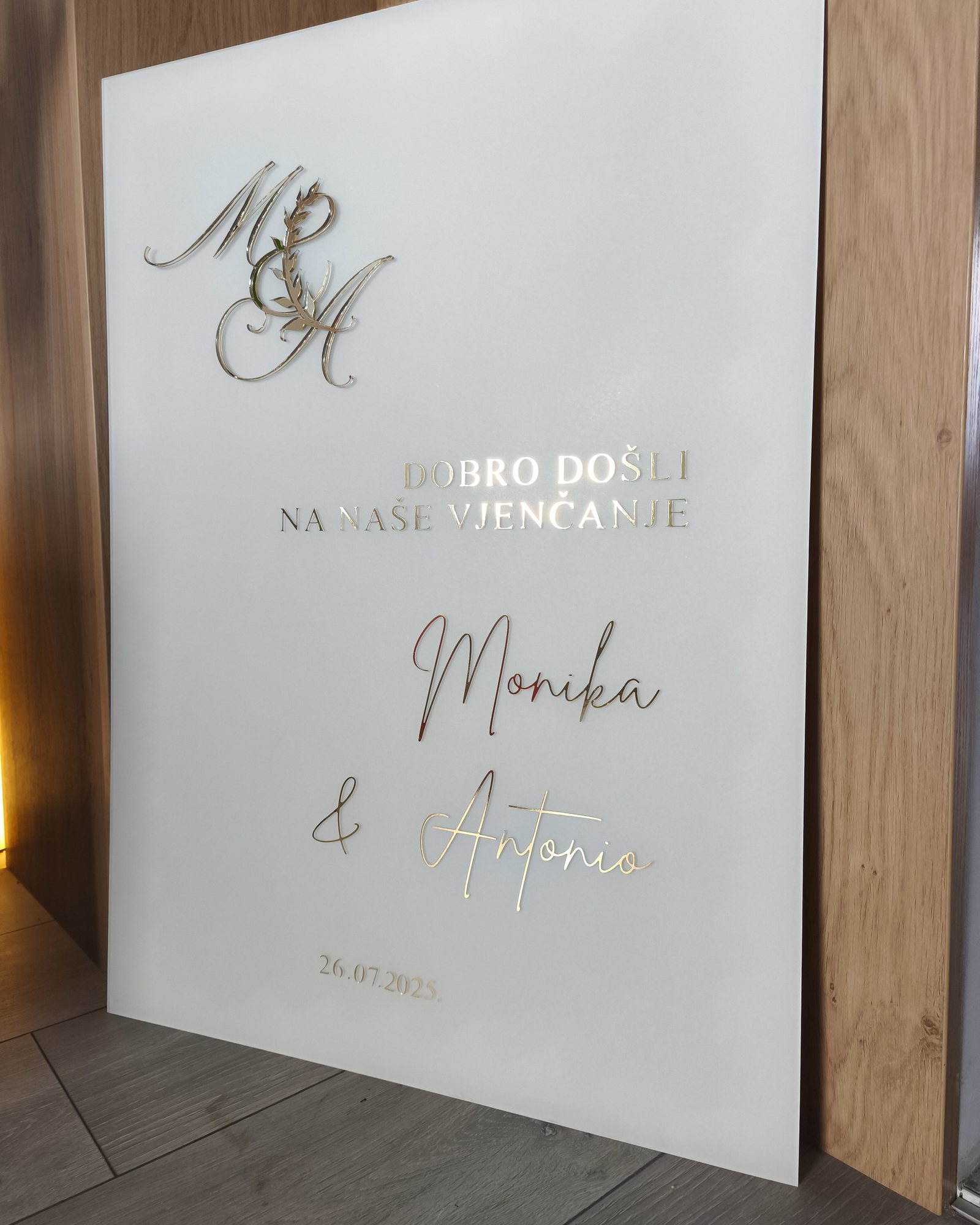 Bijela akrilna welcome ploča sa zlatnim monogramom M&A i natpisom „Dobro došli na naše vjenčanje, Monika & Antonio, 26. 7. 2025.“, naslonjena na drveni zid.