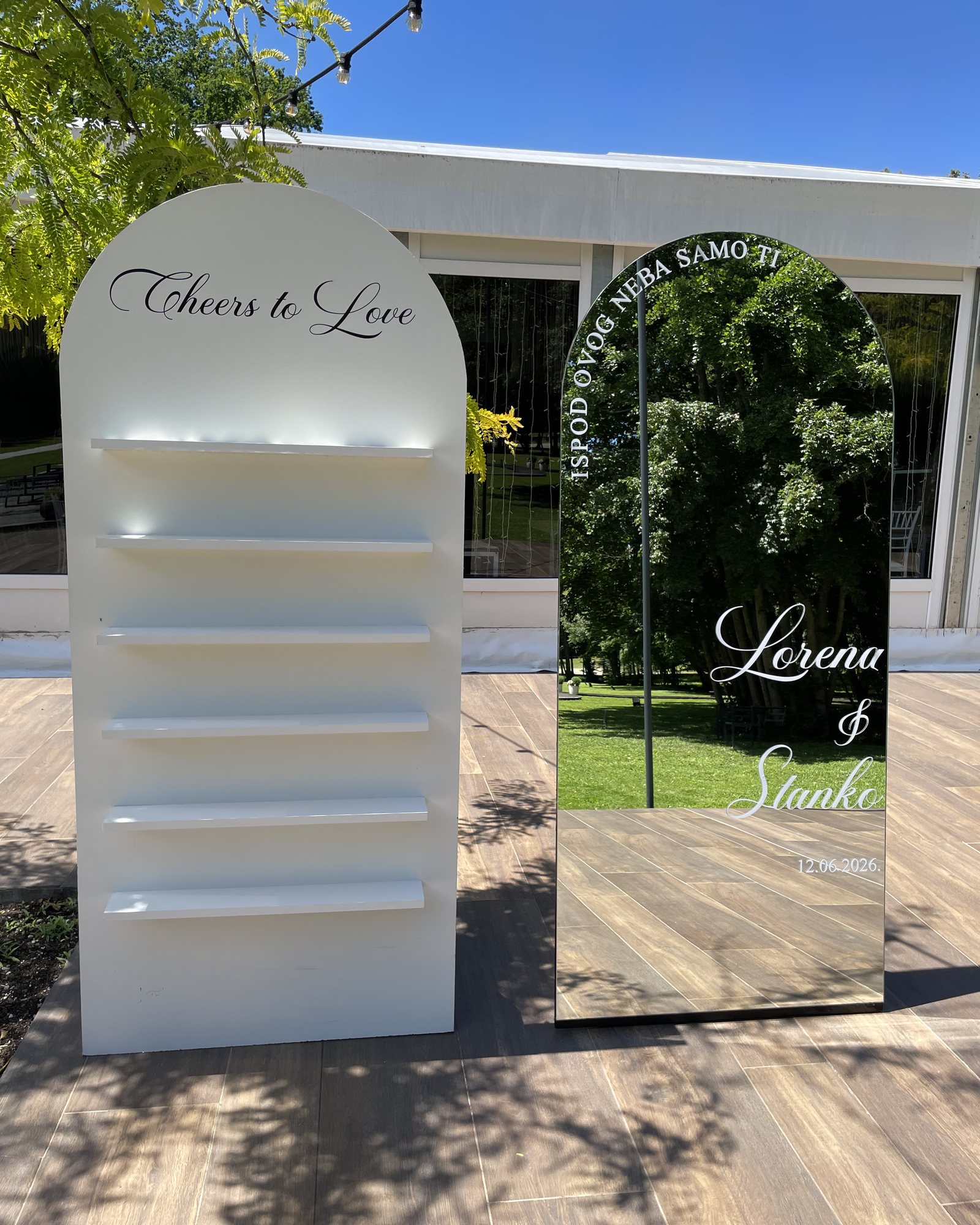 Bijela lučna polica za čaše s natpisom „Cheers to Love“ i lučno ogledalo s imenima Lorena & Stanko na terasi ispred dvorane.